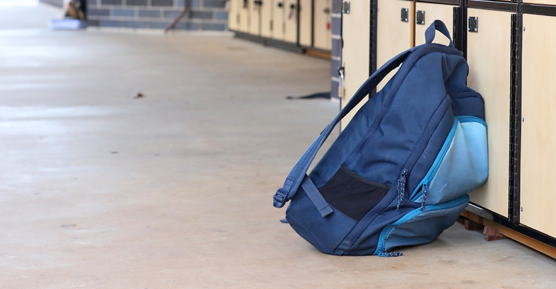 na foto, o corredor vazio de uma escola. Em primeiro plano, há uma mochila azul encostada em um armário escolar.