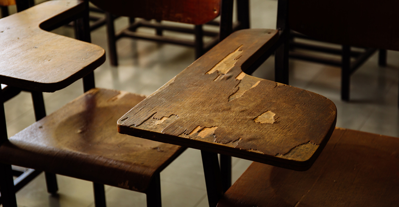a foto destaca duas carteiras escolares de madeira. A superfície de uma delas está descascada.